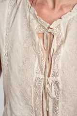 Blusa sin mangas de color beige, confeccionada en tela ligera con detalles de bordados florales frontales, encajes y cierre delantero con tiras para anudar.