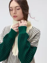 Pullover de tejido acanalado grueso con cuello alto y cierre frontal parcial. Presenta un diseño de bloques de color, con cuerpo en tono beige y mangas en contraste con rayas verticales en tonos rojizos. Incluye un detalle bordado en el pecho.