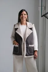 Chaleco de gamuza sintética en color gris oscuro con interior y bordes de corderito beige. Presenta solapas amplias, dos bolsillos frontales con ribetes de corderito y diseño sin cierre.