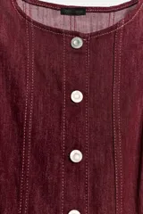 Vestido corto de denim color bordó, sin mangas, con cuello redondo, costuras pespunteadas a contratono y cierre frontal con fila de botones metálicos.