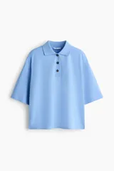 Remera polo de piqué de algodón en color celeste, con cuello acanalado, tapeta de tres botones, mangas al codo y hombros caídos. Presenta un corte holgado con bajo recto y aberturas laterales.
