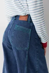 Jean de corte wide leg con tiro alto, confeccionado en denim azul oscuro. Presenta costuras verticales a lo largo de las piernas con pespuntes en color verde a contratono.