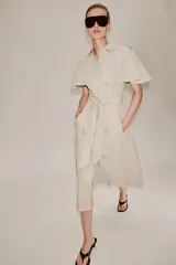 Vestido camisero midi de algodón con diseño de capa, cuello solapa, manga corta tipo capa y trabillas en los hombros. Presenta cierre frontal con botones, bolsillos delanteros y cinturón ajustable del mismo tejido con lazada. El bajo tiene un acabado en línea evasé.