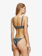 Braga de bikini tipo tanga con estampado de camuflaje en tonos grises y logo metálico ovalado en la parte delantera.