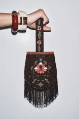 Bolso pequeño de mano con asa corta, confeccionado con abalorios negros y diseño floral bordado en tonos rosa y verde. Presenta flecos largos de abalorios en la parte inferior y cierre con botón a presión.