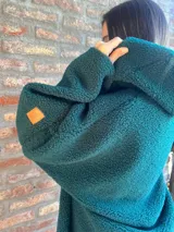 Campera de corderito color verde oscuro, con diseño holgado, cuello alto y manga larga. Presenta una etiqueta de cuero sintético en la manga.