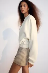 Falda pantalón de tiro bajo color beige con diseño de rayas diplomáticas verticales en tonos oscuros, con detalles de cuatro botones negros decorativos en la parte frontal y cierre lateral con cremallera oculta.
