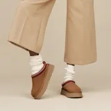 Pantuflas tipo mule UGG modelo Tazz II, confeccionadas en gamuza color tostado con plataforma alta. Presentan un detalle de costura trenzada decorativa en el borde superior en tonos rojo y beige, y el logo de la marca grabado en el lateral.