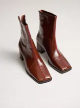 Botas de caña media color marrón, confeccionadas en cuero con acabado brillante. Presentan punta cuadrada, taco de madera y un distintivo bordado decorativo en color rojo que recorre el empeine hasta la punta.