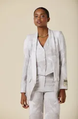 Saco de mujer con silueta estructurada, solapas anchas y cierre frontal con botones. Presenta un estampado floral sutil en tonos grisáceos y un parche decorativo con texto en la manga izquierda.
