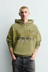 Sudadera con capucha de corte relajado en color verde oliva con efecto lavado. Presenta un bolsillo frontal tipo canguro y un estampado gráfico en el pecho con letras en tonos amarillos y negros. Cuenta con puños y bajo acanalados.