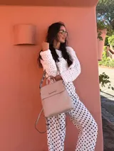 Mochila tipo cartera con tapa, confeccionada en símil cuero color beige. Presenta un diseño moderno con detalle de ojales metálicos en la parte frontal, compartimentos internos organizadores y bolsillo trasero con cierre.