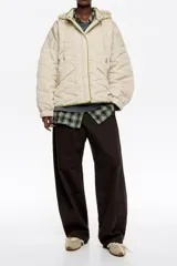 Chaquetón corto de corte línea A, confeccionado en tejido técnico acolchado color beige claro, con costuras en patrón geométrico. Posee cuello subido con capucha integrada, cierre frontal oculto con botones de presión, manga larga con puño elástico y vivos de ribete verde lima al contraste.