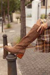 Botas de caña alta confeccionadas en cuero gamuzado color marrón. Presentan un diseño de estilo western con puntera cuadrada, taco de madera y una tira decorativa con estribo metálico desmontable en la zona del tobillo.