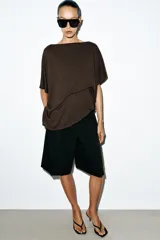 Top tipo capa blanco de punto fluido, con mezcla de viscosa, cuello bote, mangas cortas y bajo asimétrico.