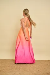 Vestido largo de satén con diseño degradado en tonos salmón y fucsia. Presenta cuello halter con detalle de nudo, falda evasé con tajo frontal pronunciado y espalda descubierta.
