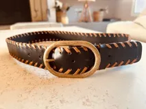 Cinturón de cuero negro con costuras decorativas color camel y hebilla ovalada de metal color bronce.