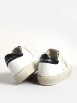 Zapatillas Golden Goose modelo Hi Star, de cuero blanco con estrella dorada con cristales Swarovski, puntera de gamuza gris y refuerzo trasero de cuero negro brillante. La suela es beige con texto manuscrito.