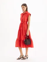Vestido camisero largo color rojo, de mangas cortas con hombros estructurados y falda escalonada con volados. Presenta cierre frontal con botones y lazo ajustable en la cintura del mismo material.
