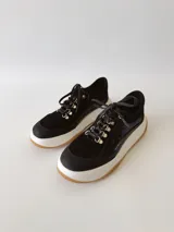 Zapatillas deportivas negras de cuero, con cordones y suela de goma blanca con base marrón.