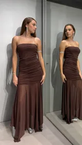 Vestido largo color chocolate, con escote strapless y diseño ajustado en el torso con frunces laterales. La falda presenta un corte evasé con una capa superior de tejido semitransparente que aporta movimiento y elegancia.