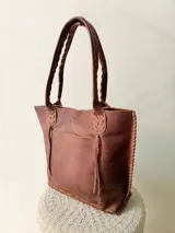 Cartera tipo shopper de cuero genuino marrón, con terminaciones artesanales trenzadas en las asas y los laterales, bolsillo frontal externo y asas largas para llevar al hombro.
