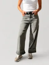 Pantalón de jean estilo palazzo, color gris claro, con corte amplio y fluido. Presenta un dobladillo ancho fijo en la botamanga, cierre frontal con botón y cremallera, y trabillas para cinturón. Incluye un cinturón negro con hebilla metálica.