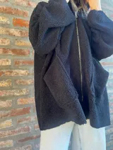 Campera de corderito color verde oscuro, con diseño holgado, cuello alto y manga larga. Presenta una etiqueta de cuero sintético en la manga.