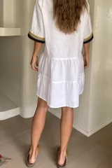 Vestido corto blanco de lino, con mangas amplias y escote en V con botones. Presenta detalles de bandas en tonos beige y negro con puntillas decorativas en el frente y en los puños. Diseño de falda con volados escalonados y silueta holgada.