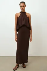 Vestido largo de color marrón oscuro con cuello halter y lazada en la nuca. Presenta un diseño con detalle de tejido drapeado efecto globo en la cintura y corte recto en la falda.