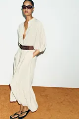 Vestido midi color crudo, con cuello camisero, mangas abullonadas por debajo del codo con puños elásticos y cierre frontal oculto con botones. Incluye bolsillos laterales y un cinturón ancho de cuero marrón oscuro con hebilla metálica.