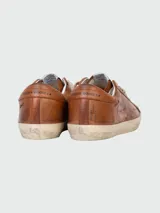 Zapatillas Golden Goose modelo Super-Star de cuero color marrón con efecto desgastado. Presentan cordones color crema, suela de goma envejecida y detalles de escritura grabada en el lateral y la puntera.