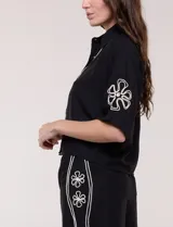 Conjunto de dos piezas negro compuesto por camisa de manga corta con cuello camisero y pantalón palazzo de tiro alto. Presenta detalles de bordados florales en contraste a lo largo de los laterales del pantalón y en las mangas.