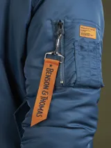Campera tipo bomber color azul, con cierre frontal, puños y cintura acanalados. Presenta un bolsillo utilitario en la manga con una cinta decorativa naranja con la inscripción Benson & Thomas.