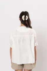 Camisa corta color blanco de corte amplio, con mangas cortas y sutiles detalles de volados a lo largo de la botonadura frontal.