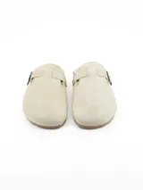 Pantufla tipo zueco en color beige, confeccionada en material tipo gamuza con interior forrado en piel sintética. Cuenta con una tira ajustable en el empeine con hebilla metálica lateral.