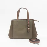 Cartera tipo tote de diseño estructurado, combinando paneles laterales de cuero sintético marrón con un panel central de tela texturizada en color verde oliva. Incluye doble asa superior, correa larga ajustable y extraíble, y un dije decorativo con cuentas de madera y piedra.