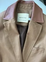 Tapado de lana color tostado con solapas clásicas y cuello en cuero sintético de tono rosado. Presenta un diseño minimalista con cierre frontal de botones y forro interior oscuro.