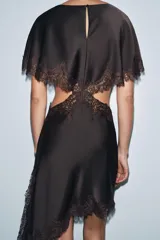 Vestido midi negro de tejido satinado con cuello redondo y manga capa. Presenta detalles de cut-out laterales adornados con encaje a tono. El bajo es asimétrico y tiene cierre en la espalda.