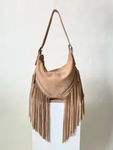 Bolso tipo hobo color tostado, hecho de cuero con flecos largos en el frente y dos bolsillos con cierre. Tiene una manija regulable con hebillas metálicas.