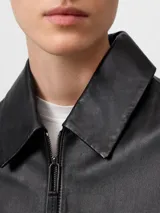 Chaqueta negra de piel ovina con diseño desestructurado, cuello solapa, cierre frontal con cremallera y bolsillos de vivo inclinados. Presenta una capelina en la espalda y mangas largas.