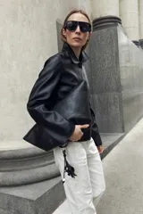 Chaqueta de eco cuero negro con cuello camisero, cierre frontal oculto y mangas largas con puños rectos.