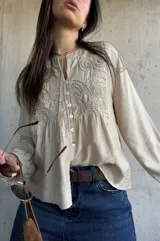Camisa beige de manga larga con detalles bordados en el frente, cuello redondo y cierre frontal con botones. Las mangas presentan puños con volados.