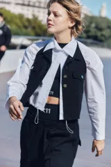 Chaleco de denim negro con escote halter, cuello camisero y cierre frontal con botones metálicos. Presenta dos bolsillos frontales con solapa y diseño sin mangas.