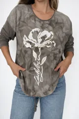 Remera blanca de línea italiana, confeccionada en algodón y viscosa, con calce holgado y cuello redondo. Presenta un estampado frontal de una flor en tonos oscuros con detalles dorados y la frase "Ladybee". Incluye tiras regulables en la parte inferior para ajustar el largo.
