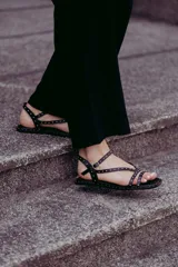 Sandalias planas de cuero negro con múltiples tiras finas adornadas con tachas doradas. Cuentan con cierre de hebilla en el tobillo.