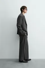 Pantalón de vestir gris oscuro, corte ancho (flare fit) y tiro alto, con bolsillos laterales y falsos bolsillos de vivo en la parte trasera. Presenta un cierre frontal con cremallera y gancho oculto, y botones a presión ocultos en el bajo.