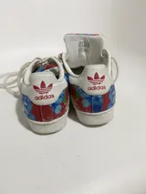 Championes Adidas modelo Superstar, con diseño estampado floral en tonos azules, rojos y amarillos sobre base de tela, con la clásica puntera de goma blanca y cordones blancos.