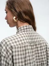 Camisa de corte clásico con estampado de cuadros vichy en tonos azul y blanco. Presenta cuello camisero, cierre frontal con botones ocultos y mangas largas con puños abotonados. Incluye un detalle de broche metálico dorado en el cuello.