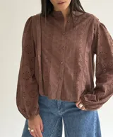 Blusa de brodery color blanco, con cuello redondo, mangas largas abullonadas con puños elásticos y cierre frontal con botones forrados. Presenta detalles de puntillas verticales en el frente.